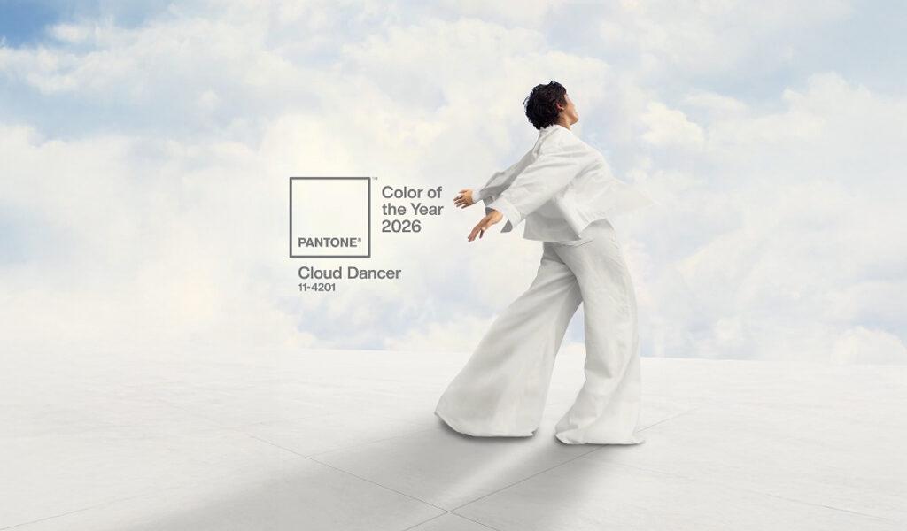Une personne habillée tout en blanc danse avec les bras ouverts devant un ciel nuageux, accompagnée du logo Pantone "Color of the Year 2026 - Cloud Dancer 11-4201".