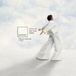 Une personne habillée tout en blanc danse avec les bras ouverts devant un ciel nuageux, accompagnée du logo Pantone "Color of the Year 2026 - Cloud Dancer 11-4201".