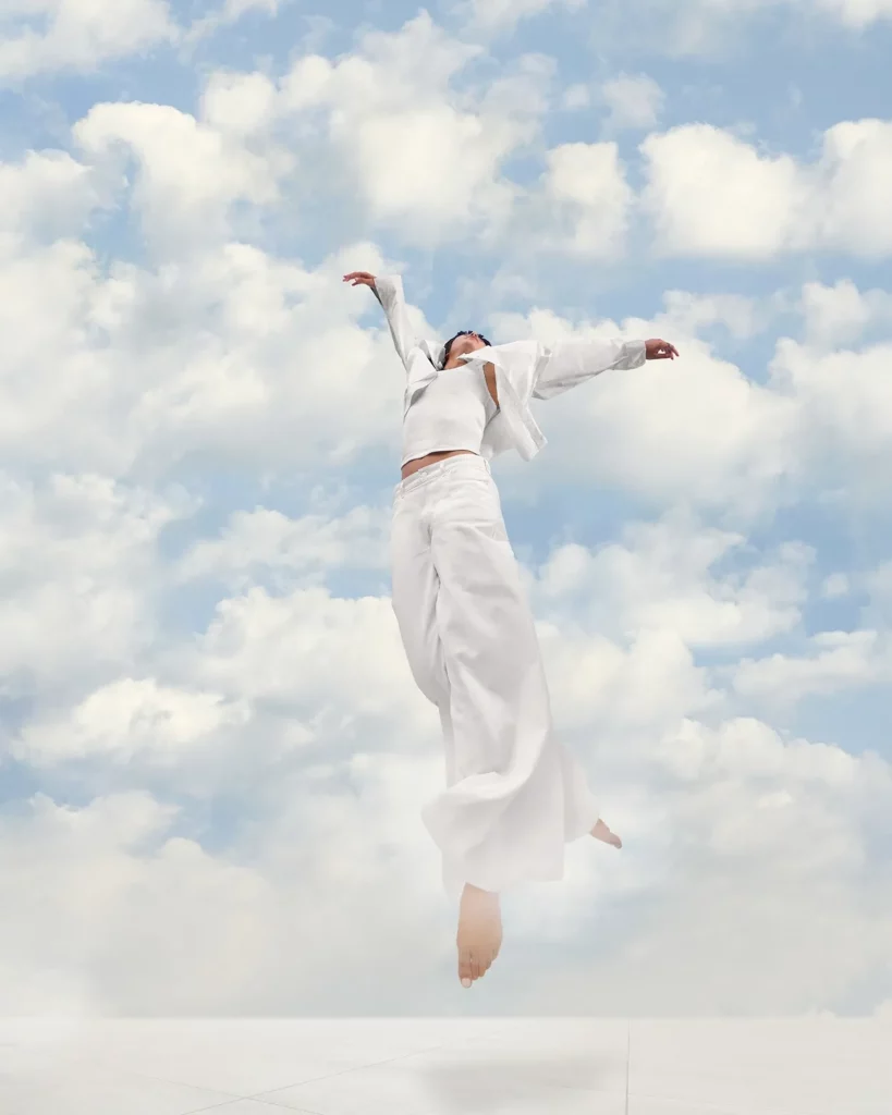 Une personne habillée tout en blanc danse avec les bras ouverts devant un ciel nuageux